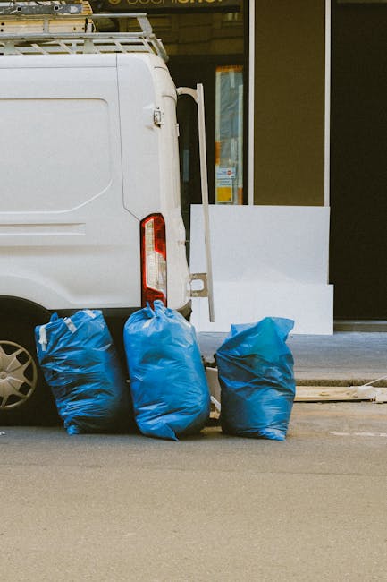A collection of black garbage bags, cardboard boxes, and discarded packaging materials piled in front of a red metal door mounted in a textured, off-white wall. The door has multiple graffiti tags in 