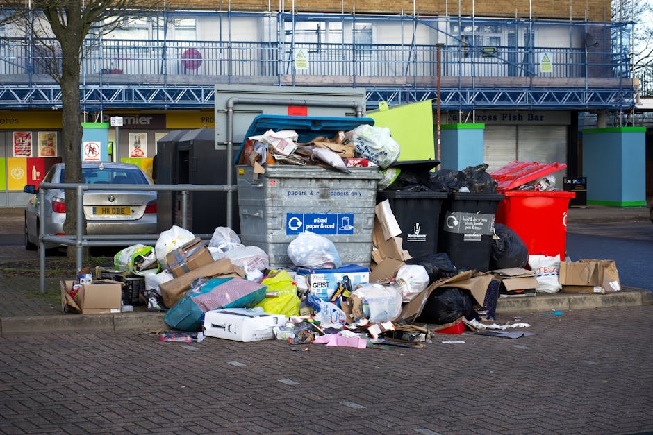 A man wearing a dark red jacket and black trousers is standing with his back to the camera, operating a large, open green waste collection bin on a busy street or marketplace environment. In the foreg