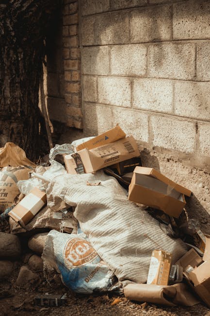 Close-up of a tightly knotted black plastic rubbish bag made of glossy, flexible material, with visible creases and folds, positioned in the foreground against a plain, light-colored wall. The bag app