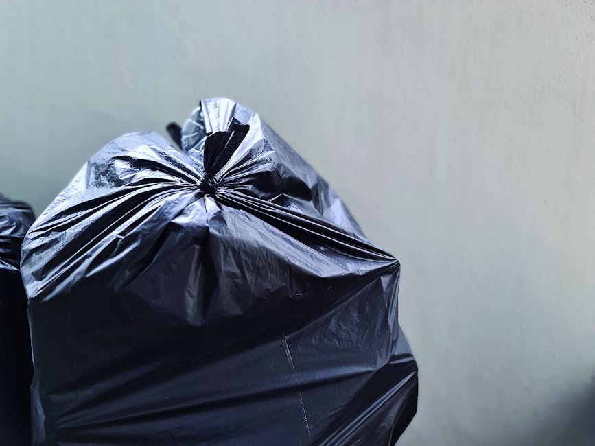 An outdoor scene showing a large pile of mixed rubbish and waste materials in a disorganized heap. The debris includes plastic bags, cardboard boxes, broken furniture, metal pipes, and discarded house